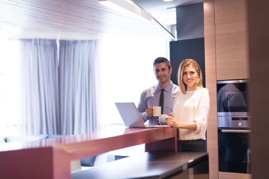 A young couple is preparing for the job and using a laptop. The man drinks coffee while the woman eats breakfast at luxury home together, looking at screen, smiling.