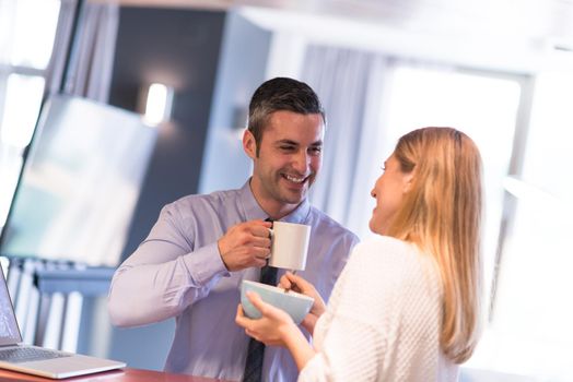 A young couple is preparing for the job and using a laptop. The man drinks coffee while the woman eats breakfast at luxury home together, looking at screen, smiling.