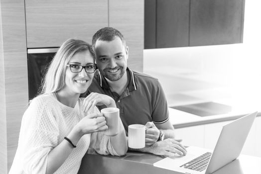 Young couple drinking coffee and using laptop computer at luxury home together, looking at screen, smiling.