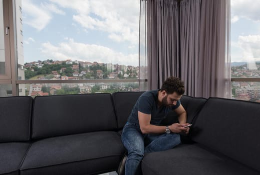 young man sitting on sofa and using a mobile phone  near the window at home