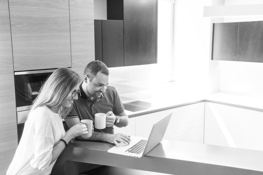 Young couple drinking coffee and using laptop computer at luxury home together, looking at screen, smiling.