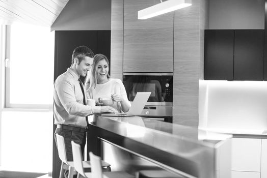 A young couple is preparing for the job and using a laptop. The man drinks coffee while the woman eats breakfast at luxury home together, looking at screen, smiling.