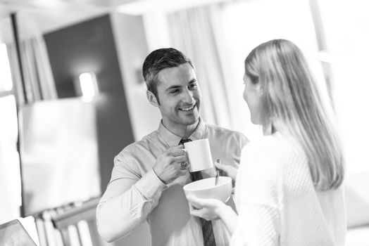 A young couple is preparing for the job and using a laptop. The man drinks coffee while the woman eats breakfast at luxury home together, looking at screen, smiling.