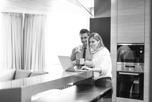 A young couple is preparing for the job and using a laptop. The man drinks coffee while the woman eats breakfast at luxury home together, looking at screen, smiling.