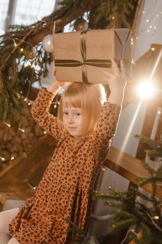 A little blonde girl is sitting on a wooden staircase in a Scandinavian interior decorated in a New Year's style. A child holds a Christmas gift in a craft package.