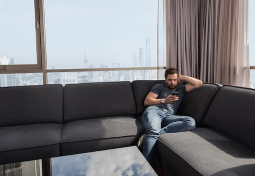 young man sitting on sofa and using a mobile phone  near the window at home