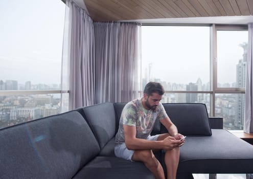 young man sitting on sofa and using a mobile phone  near the window at home