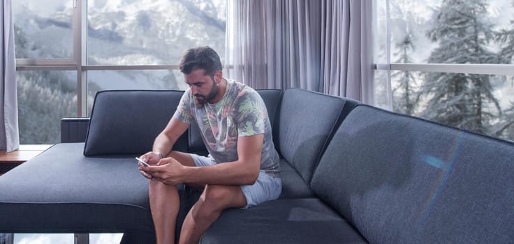 young man sitting on sofa and using a mobile phone  near the window at home