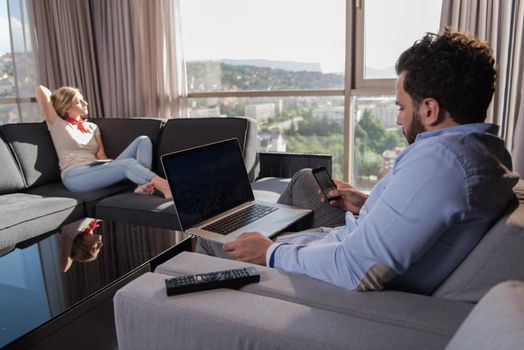 Young couple relaxing at home using tablet and laptop computers reading near the window on the sofa couch.