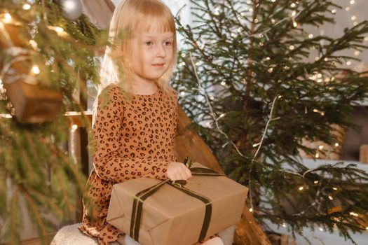 A little blonde girl is sitting on a wooden staircase in a Scandinavian interior decorated in a New Year's style. A child holds a Christmas gift in a craft package.