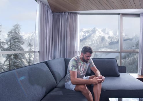 young man sitting on sofa and using a mobile phone  near the window at home