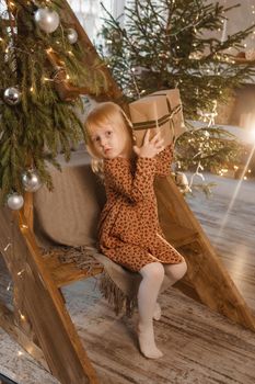 A little blonde girl is sitting on a wooden staircase in a Scandinavian interior decorated in a New Year's style. A child holds a Christmas gift in a craft package.