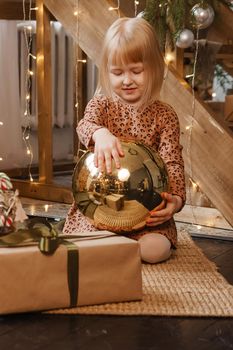 A little blonde girl is sitting next to a wooden staircase in a Scandinavian interior decorated in a New Year's style. A child holds a golden Christmas ball.