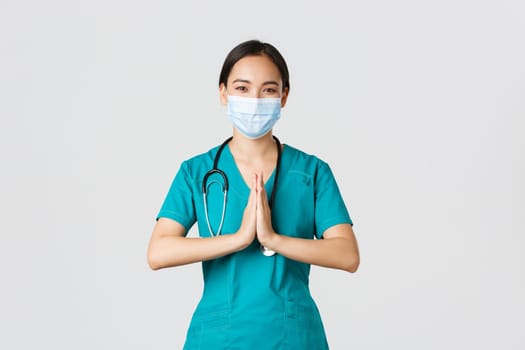 Covid-19, coronavirus disease, healthcare workers concept. Hopeful smiling asian female physician, doctor in medical mask and scrubs hold hands in praying gesture, thanking, white background.