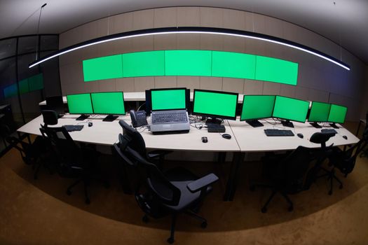 Empty interior of big modern security system control room with blank green screens, workstation with multiple displays, monitoring room with at security data center  Empty office, desk, and chairs at a main CCTV security data center