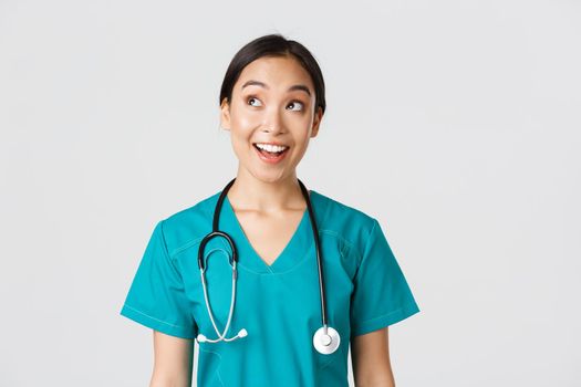 Healthcare workers, preventing virus, quarantine campaign concept. Enthusiastic smiling asian doctor in scrubs, nurse looking upper left corner with amused face, standing white background.