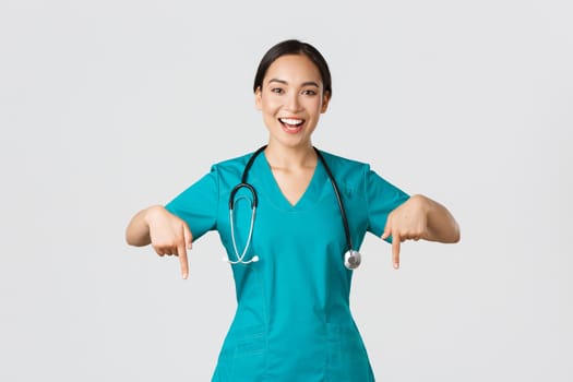 Covid-19, healthcare workers, pandemic concept. Happy smiling beautiful asian nurse, female doctor in scrubs pointing down, inviting for checkup, showing hospital advertisement, white background.