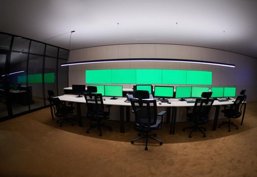 Empty interior of big modern security system control room with blank green screens, workstation with multiple displays, monitoring room with at security data center  Empty office, desk, and chairs at a main CCTV security data center