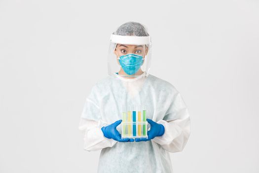 Covid-19, coronavirus disease, healthcare workers concept. Surprised asian female doctor, tech lab in personal protective equipment holding test-tubes with vaccine or analyses, white background.