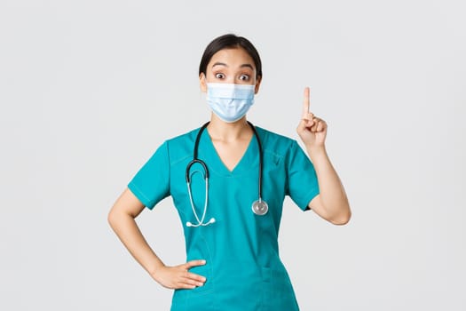 Covid-19, coronavirus disease, healthcare workers concept. Excited asian female doctor, physician in medical mask and scrubs have suggestion, raising index finger eureka gesture, white background.