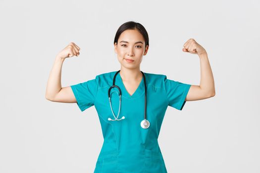 Covid-19, healthcare workers and preventing virus concept. Confident professional asian female doctor, nurse in scrubs smiling sassy and flex biceps, being strong, show-off muscles.