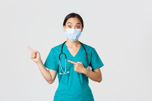 Covid-19, coronavirus disease, healthcare workers concept. Surprised and interested asian female doctor, physician in medical mask and scrubs pointing fingers upper left corner, white background.
