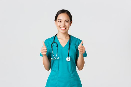 Covid-19, healthcare workers, pandemic concept. Professional confident smiling doctor, female physician in scrubs showing thumbs-up with assured expression, ensure all good, perfect or excellent.