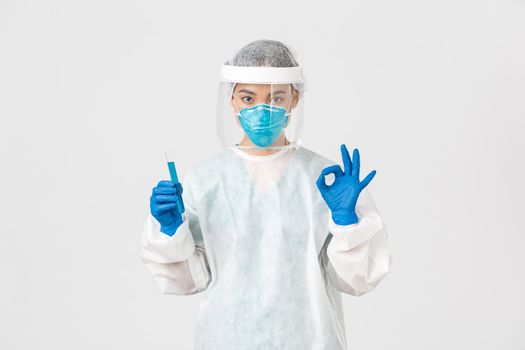 Covid-19, coronavirus disease, healthcare workers concept. Serious asian female doctor, professional researcher in personal protective equipment showing okay gesture, holding test-tube with vaccine.