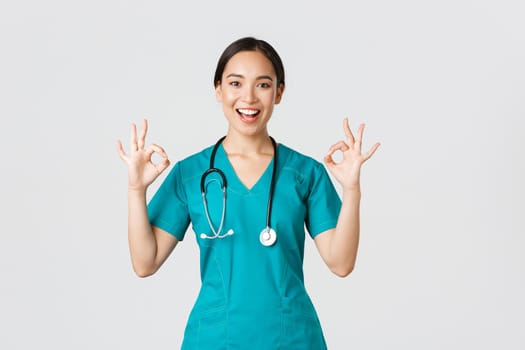 Covid-19, healthcare workers, pandemic concept. Professional confident asian female doctor, intern in scrubs assure patients everything good, showing okay gesture satisfied, smiling pleased.