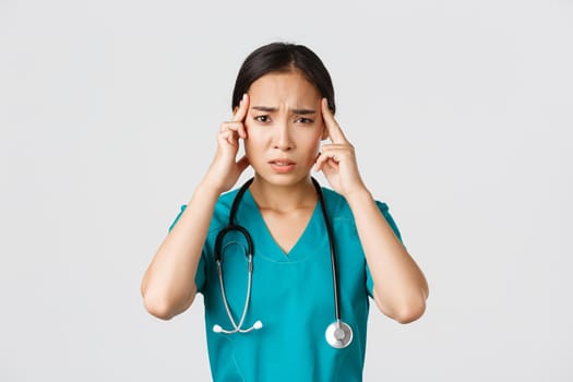 Healthcare workers, preventing virus, quarantine campaign concept. Exhausted asian female doctor feeling dizzy, touching head and grimacing from headache, standing white background.