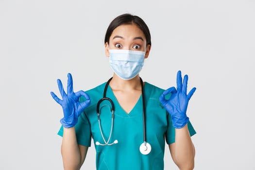 Covid-19, coronavirus disease, healthcare workers concept. Excited and impressed asian doctor, nurse in medical mask and rubber gloves showing okay gesture, approve or praise nice work.