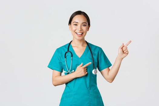 Covid-19, healthcare workers, pandemic concept. Smiling cheerful asian female doctor, therapist in scrubs pointing fingers upper right corner, inviting for checkup, recommend vaccinate.