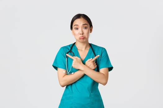 Covid-19, healthcare workers, pandemic concept. Indecisive and clueless asian female nurse, intern dont know what choose, pointing fingers sideways and shrugging unaware, white background.