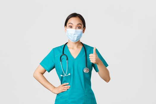 Covid-19, coronavirus disease, healthcare workers concept. Amused and impressed asian female doctor, nurse in scrubs and medical mask, show thumb-up in approval, agree or compliment work.