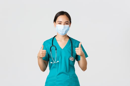 Covid-19, coronavirus disease, healthcare workers concept. Confident, smiling asian doctor, female nurse in scrubs and medical mask profive good quality service, show thumbs-up in approval.
