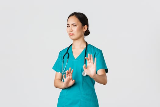 Covid-19, healthcare workers, pandemic concept. Reluctant funny asian nurse, doctor avoiding someone, step away and show stop gesture, grimacing or cringe as rejecting awful offer, refuse.