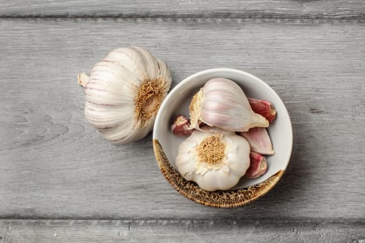 Table top view on garlic bulbs and peeled cloves in small ceramic bowl placed on gray wood desk.