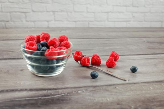 Silver spoon full of raspberries and blueberries, some spilled on gray wood desk, with out of focus bowl full of more berries on the side.