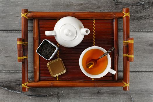 Table top view on white ceramic teapot, cup of hot black tea with silver spoon and opened dose with loose leaf tea, served on bamboo tray placed on gray wood desk.