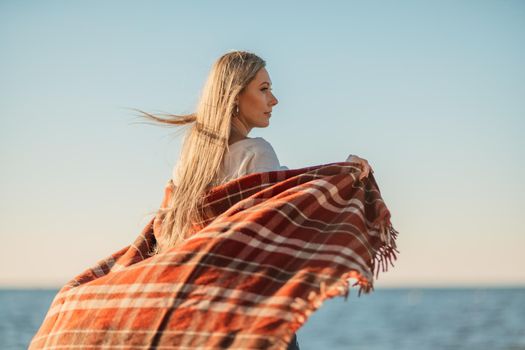 Attractive blonde Caucasian woman enjoying time on the beach at sunset, walking in a blanket and looking to the side, with the sunset sky and sea in the background. Beach vacation.