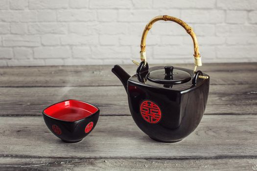 Black teapot and square cup full of hot tea with steam, with red Chinese seal meaning "Double Happiness", placed on gray wood table.