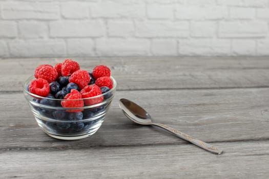 Small glass bowl full of blueberries with raspberries on top, with silver spoon next to it suggests its ready to eat, placed on gray wood desk.