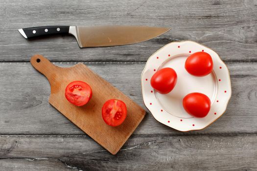 Table top view on three tomatoes on white porcelain plate with red dots, cutting board with tomato cut to half next to it, chef knife also laying on the gray wood desk.