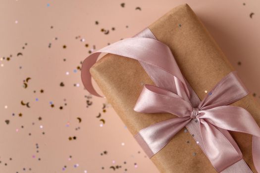 Birthday or New Year's eve present in kraft paper with pink ribbon on a soft beige background with glitter and confetti. Xmas composition. Flat lay. Happy holidays celebration and giving love concept