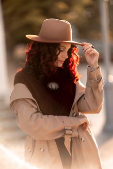 Outdoor fashion portrait of young elegant fashionable brunette woman, model in stylish hat, choker and light raincoat posing at sunset in European city