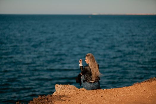 A blonde girl in a stylish black leather jacket is sitting with her back to the seashore