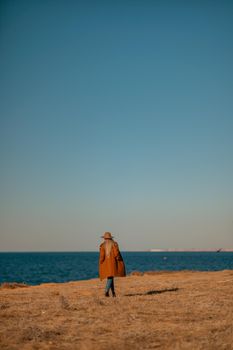 A woman walking along the coast near the sea. An elegant lady in a brown coat and a hat with fashionable makeup walks on the seashore.