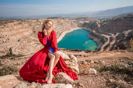 A beautiful girl in a red long dress, Sits on a rock high above the lake in the afternoon. Against the background of the blue sky and the lake in the shape of a heart