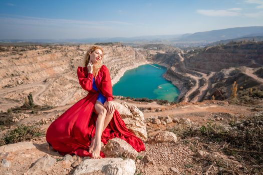 A beautiful girl in a red long dress, Sits on a rock high above the lake in the afternoon. Against the background of the blue sky and the lake in the shape of a heart