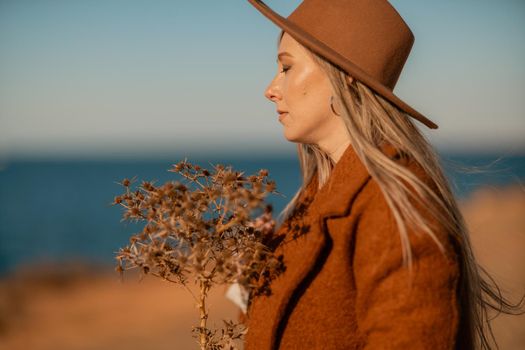 A woman walking along the coast near the sea. An elegant lady in a brown coat and a hat with fashionable makeup walks on the seashore.